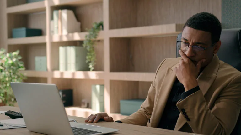 Businessman sitting at a desk with his hand over his mouth while looking at a laptop
